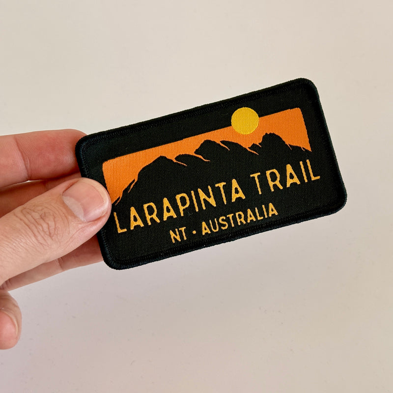 Load image into Gallery viewer, Hand holding a black iron-on patch with 'Larapinta Trail NT Australia' design on a beige background
